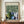May include: Framed poster with William Morris Gallery exhibition text at top, featuring green botanical pattern with cream flowers and leaves in upper half, abstract painted elements in blues, greens, and whites below, quote at bottom. Wooden frame on light wood credenza beside white ceramic vase and stacked books against cream wainscoted wall.