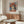 May include: Framed art print of woman wearing wide-brimmed red hat and red top against blue background, displayed above wooden sideboard with glass doors. Modern living room with curved white chair, oval glass coffee table, floor lamp, and table lamp. Sunlight creates shadow patterns on polished floor.