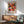 May include: Yayoi Kusama exhibition poster from 1984 featuring colorful polka dot patterns in pink, orange, and coral tones, displayed in wooden frame above mid-century modern sideboard. Green upholstered armchair with wooden arms sits nearby on hardwood floor, white table lamp creates warm lighting, sunlight streams across white wall creating geometric shadows.