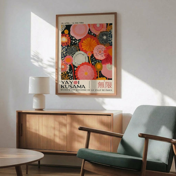 May include: Yayoi Kusama exhibition poster from 1984 featuring colorful polka dot patterns in pink, orange, and coral tones, displayed in wooden frame above mid-century modern sideboard. Green upholstered armchair with wooden arms sits nearby on hardwood floor, white table lamp creates warm lighting, sunlight streams across white wall creating geometric shadows.