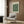 May include: Framed vintage botanical poster with green floral pattern mounted on wood paneled wall above beige upholstered armchair with geometric patterned pillow. Modern brass floor lamp and dark round side table with open book complete the neutral-toned reading nook setting.