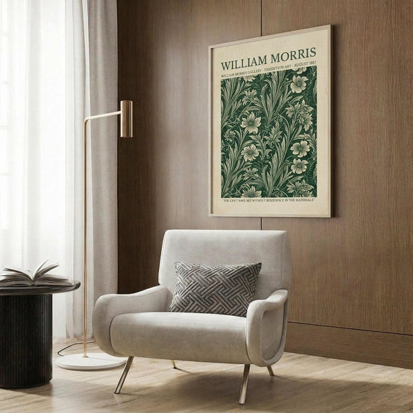 May include: Framed vintage botanical poster with green floral pattern mounted on wood paneled wall above beige upholstered armchair with geometric patterned pillow. Modern brass floor lamp and dark round side table with open book complete the neutral-toned reading nook setting.