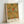 May include: Framed poster featuring William Morris floral pattern with golden yellow carnations, red flowers, and green foliage on cream background. Light wood frame leans against white wall corner, showing exhibition art print with designer's name at bottom.
