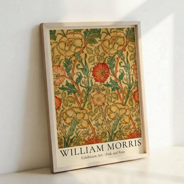 May include: Framed poster featuring William Morris floral pattern with golden yellow carnations, red flowers, and green foliage on cream background. Light wood frame leans against white wall corner, showing exhibition art print with designer's name at bottom.