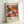 Colorful exhibition poster featuring vibrant circular and radial patterns in pink, orange, yellow, and black with white dots scattered throughout. Text shows dates "28. APRIL - 14. MAI 1984" at top and "YAYOI KUSAMA" with Japanese characters and "MUSEE D'ART MODERNE DE LA VILLE DE PARIS" at bottom. The artwork displays overlapping floral and geometric shapes creating a dynamic, psychedelic composition. Poster is displayed in a light wooden frame leaning against a white wall