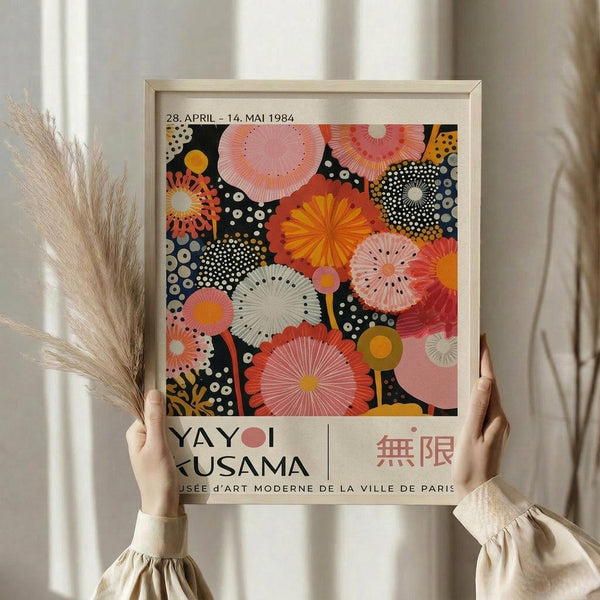 May include: Person holding framed poster featuring colorful abstract circular patterns in pink, orange, yellow and black with dotted details. Light wood frame against neutral curtained background with dried pampas grass visible. Exhibition poster shows dates 28 April - 14 Mai 1984 and Musée d'Art Moderne de la Ville de Paris text below vibrant radiating botanical-inspired design.