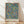 May include: Framed botanical art print leaning against white wall on light wood floor. Features intricate pattern of fruits, pomegranates, and leafy branches on soft blue-gray background with yellow, orange, red, and green details. Natural wood frame with artist name text below artwork. Soft natural lighting creates gentle shadows on textured white wall.