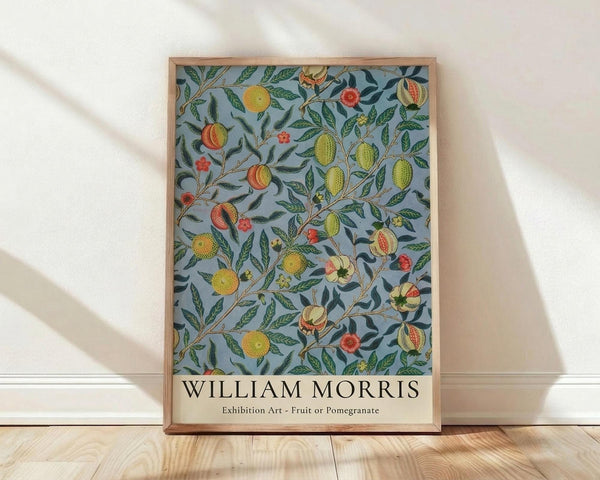 May include: Framed botanical art print leaning against white wall on light wood floor. Features intricate pattern of fruits, pomegranates, and leafy branches on soft blue-gray background with yellow, orange, red, and green details. Natural wood frame with artist name text below artwork. Soft natural lighting creates gentle shadows on textured white wall.
