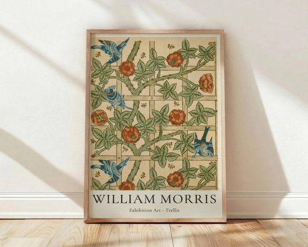 May include: Framed botanical poster leaning against white wall on light wood floor. Features William Morris's Trellis pattern with blue birds perched among climbing rose vines, orange-red blooms, and green foliage arranged in grid-like trellis design on cream background. Natural wood frame with soft window light casting shadows across white textile surface.