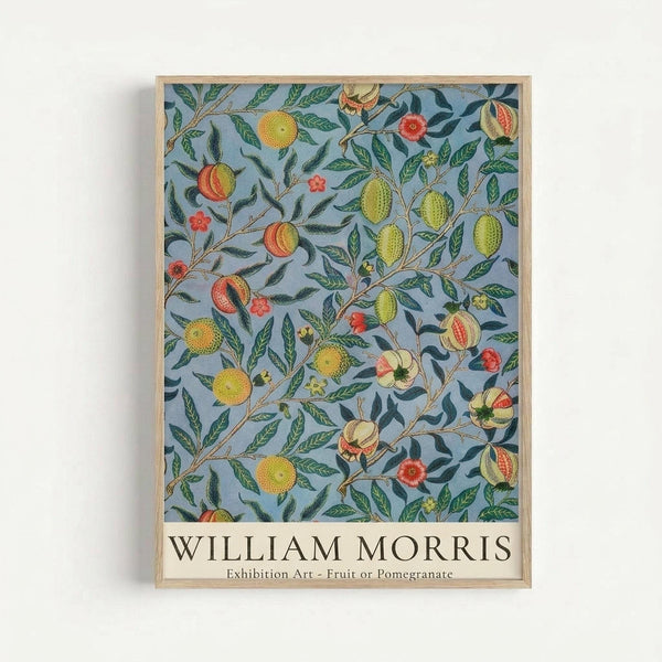 A framed botanical art print featuring William Morris's intricate pattern of fruits and foliage on a soft blue-gray background. The design shows detailed illustrations of oranges, lemons, pomegranates, and red flowers intertwined with green leaves and brown branches. The artwork is displayed in a natural wood frame against a white wall, with "WILLIAM MORRIS" and "Exhibition Art - Fruit or Pomegranate" text at the bottom.