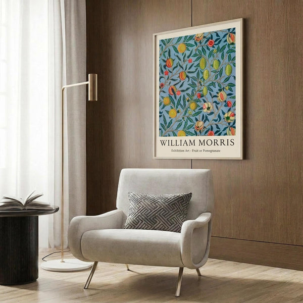 May include: Framed botanical poster with blue background featuring colorful fruits and green foliage in William Morris style, displayed above beige upholstered chair with geometric pillow. Modern living room setting with brass floor lamp, dark round side table with open book, and wood paneled wall. Natural lighting from window with sheer curtains creates warm, sophisticated interior design scene.