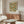 May include: Framed botanical poster with blue birds, orange flowers, and green foliage on cream background displayed above walnut wood credenza in neutral living room. White curved chair and glass coffee table visible in foreground with natural sunlight casting shadows across scene.