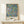 May include: A framed botanical art print featuring various fruits including pomegranates, citrus, and other round fruits with green leaves on a soft blue background, displayed on a light wood console table beside a small wrapped gift with natural twine and greenery sprig against a cream wall.