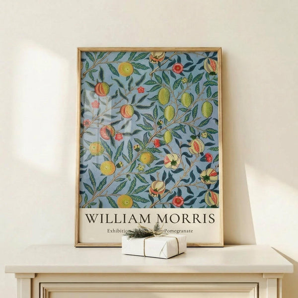 May include: A framed botanical art print featuring various fruits including pomegranates, citrus, and other round fruits with green leaves on a soft blue background, displayed on a light wood console table beside a small wrapped gift with natural twine and greenery sprig against a cream wall.