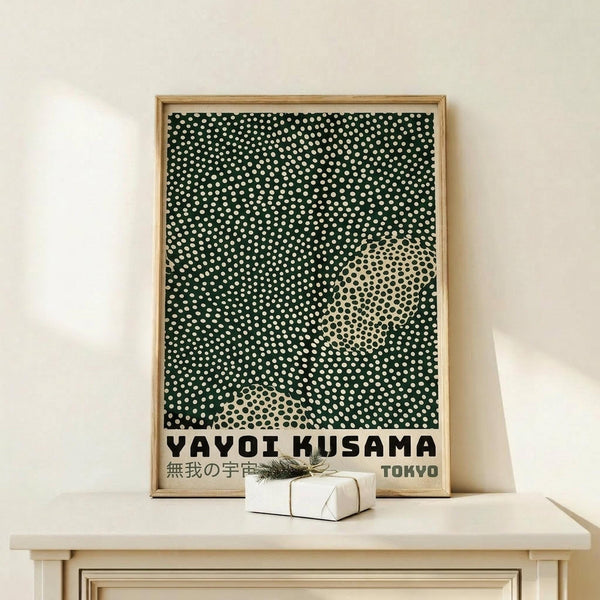 May include: Framed poster with black background and white polka dot pattern creating abstract shapes, featuring "YAYOI KUSAMA" and "TOKYO" text at bottom with Japanese characters. Natural wood frame displayed on light wooden surface against cream wall, with small white wrapped gift box beside it.