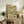 May include: Framed botanical print featuring blue birds, red flowers, and green foliage in a trellis pattern displayed on a rustic wooden sideboard beside a fern plant, books, and ceramic vase in a neutral-toned living room with beige upholstered furniture and warm natural lighting.