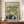 May include: Hands holding a large framed William Morris exhibition poster featuring vintage botanical wallpaper pattern with cream flowers and green foliage, displayed on wooden sideboard next to white ceramic vase and books in elegant room with white wainscoting and cream walls