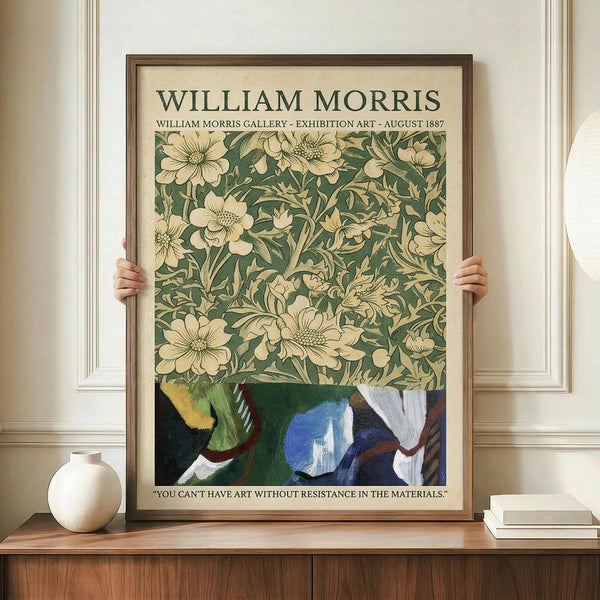 May include: Hands holding a large framed William Morris exhibition poster featuring vintage botanical wallpaper pattern with cream flowers and green foliage, displayed on wooden sideboard next to white ceramic vase and books in elegant room with white wainscoting and cream walls