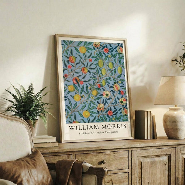 May include: Framed botanical poster with blue background featuring colorful fruits, flowers and green foliage in decorative pattern, displayed on rustic wooden sideboard alongside fern plant, ceramic vase, stacked books and cream upholstered chair in neutral-toned living room setting.