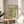 May include: Framed botanical poster with green and cream floral pattern displayed on round wooden side table next to beige upholstered chair. Natural sunlight streams across neutral-toned room with white walls and curtains. Plant leaves visible in corner. Poster features vintage-style William Morris exhibition text above densely packed flowers and foliage design in sage green and ivory tones within thin wooden frame.