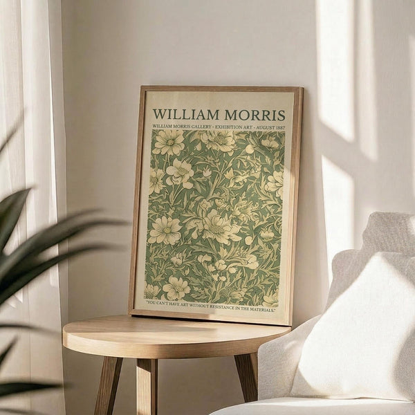 May include: Framed botanical poster with green and cream floral pattern displayed on round wooden side table next to beige upholstered chair. Natural sunlight streams across neutral-toned room with white walls and curtains. Plant leaves visible in corner. Poster features vintage-style William Morris exhibition text above densely packed flowers and foliage design in sage green and ivory tones within thin wooden frame.