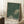 May include: Framed art print leaning against white wall showing green background with white polka dots and cream-colored circular shapes, featuring "YAYOI KUSAMA" and Japanese text at bottom with "TOKYO" underneath. Natural wood frame, positioned on white textured rug next to wooden side table with white coffee cup. Cozy interior setting with soft natural lighting and beige throw blanket visible.