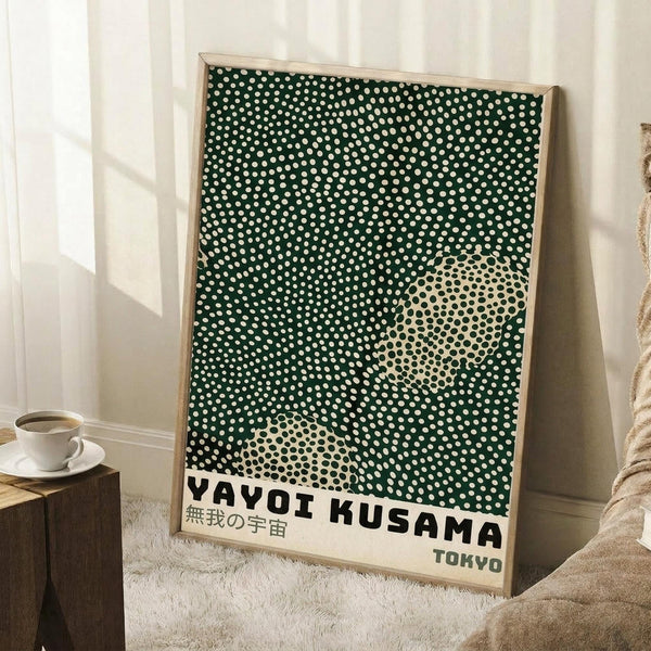 May include: Framed art print leaning against white wall showing green background with white polka dots and cream-colored circular shapes, featuring "YAYOI KUSAMA" and Japanese text at bottom with "TOKYO" underneath. Natural wood frame, positioned on white textured rug next to wooden side table with white coffee cup. Cozy interior setting with soft natural lighting and beige throw blanket visible.