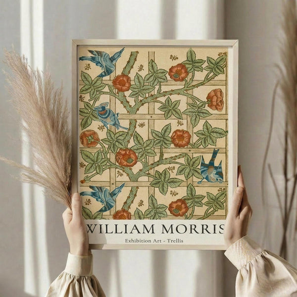 May include: Hands holding a framed botanical art print featuring blue birds, orange flowers, and green vines in William Morris style against a trellis pattern. Person wears cream ruffled sleeves, holds dried pampas grass, standing by white curtained window in soft natural lighting.