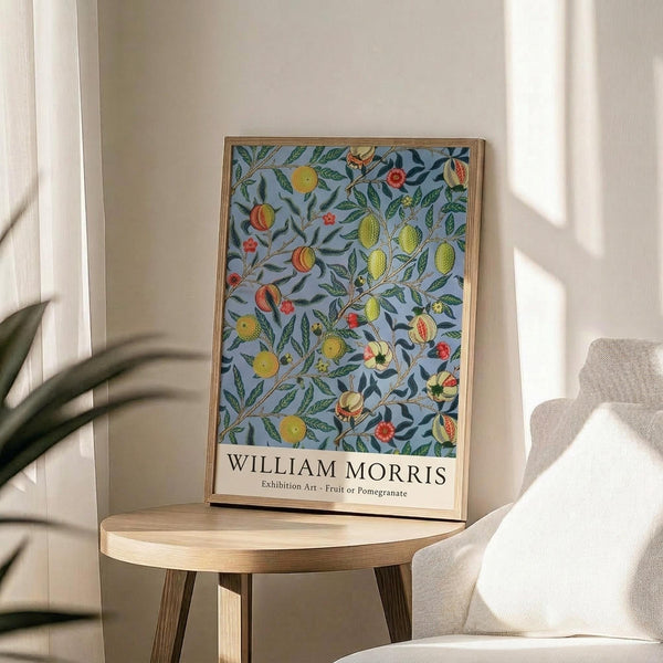 May include: Framed botanical poster with blue-grey background featuring colorful fruit and foliage pattern in William Morris style, displayed on round wooden side table next to beige upholstered chair. Natural sunlight creates soft shadows on white wall. Green plant leaves visible in corner. Artwork shows yellow, orange and red fruits with green leaves in decorative Arts and Crafts design.