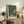 May include: Framed art print with black background and white polka dots forming circular patterns, text reading "YAYOI KUSAMA" and Japanese characters below, "TOKYO" in corner. Light wood frame leaning against white wall on rustic wooden sideboard. Scene includes cream upholstered chair, brown leather pillow, potted fern, ceramic vase, and stacked books in warm, naturally lit living room setting.