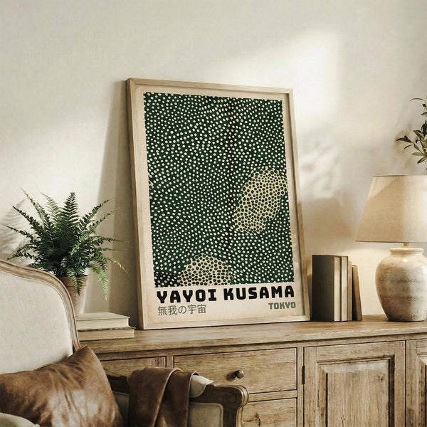 May include: Framed art print with black background and white polka dots forming circular patterns, text reading "YAYOI KUSAMA" and Japanese characters below, "TOKYO" in corner. Light wood frame leaning against white wall on rustic wooden sideboard. Scene includes cream upholstered chair, brown leather pillow, potted fern, ceramic vase, and stacked books in warm, naturally lit living room setting.