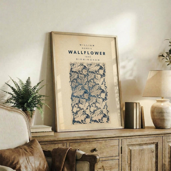 May include: Framed vintage botanical wallpaper design poster with blue floral vine pattern on cream background, displayed on rustic wooden sideboard alongside fern plant, ceramic vase, stacked books, and brown leather armchair in warm neutral living room setting