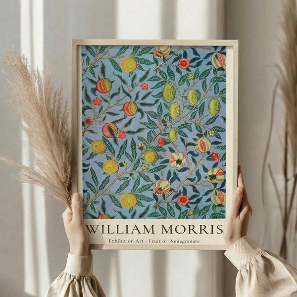 May include: Hands holding a framed botanical poster featuring colorful fruits and foliage on a light blue background in an ornate William Morris style pattern. The artwork shows oranges, lemons, pomegranates and green leaves arranged in flowing vines. Natural wood frame against white curtains with dried pampas grass visible to the left. Person wearing cream colored clothing with ruffled sleeves.