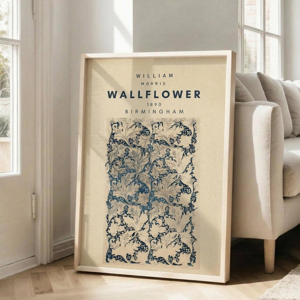 May include: Framed vintage botanical wallpaper pattern poster in cream and navy blue, featuring intricate floral and vine motifs in William Morris style. Light wood frame leans against white wall next to beige upholstered chair in bright, airy room with tall windows and natural lighting.