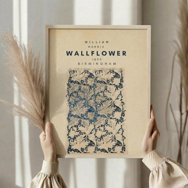May include: Hands holding a framed vintage botanical wallpaper pattern print in cream and navy blue, featuring intricate flowing floral and vine motifs. The wooden frame sits against soft beige curtains with dried pampas grass visible to the left. Person wearing a cream-colored blouse with gathered sleeves displays the artwork in natural window lighting.