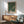 a living room with a framed poster of a green and white pattern hanging on the wall above a wooden cabinet. The poster is titled "Vavoi Kusama" and "T Tokyo", suggesting it is a piece of artwork or design related to the artist Vavoi Kusama and Tokyo. The room has a wooden floor, a green armchair, and a wooden coffee table, creating a cozy and inviting atmosphere.