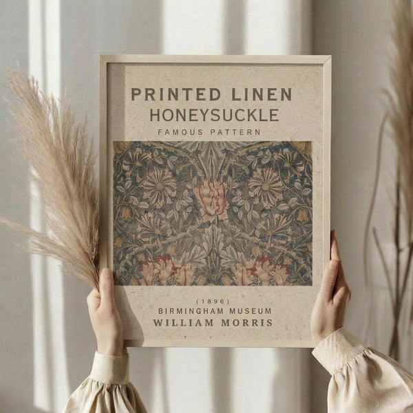 May include: Hands holding a framed vintage botanical print featuring William Morris's honeysuckle pattern from 1896. The artwork displays intricate floral motifs with pink blooms and detailed foliage in muted tones against a cream background. Light wood frame complements the neutral aesthetic, with dried pampas grass and soft lighting visible in the minimalist interior setting.