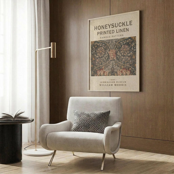 May include: Vintage botanical poster featuring William Morris honeysuckle pattern in muted greens and pinks, displayed in light wood frame on wood-paneled wall. Modern beige armchair with geometric pillow sits below artwork. Brass floor lamp and round side table with open book complete the neutral-toned reading nook with natural lighting from window.