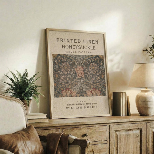 May include: Framed botanical poster with intricate honeysuckle pattern in muted greens and pinks, displayed on rustic wooden sideboard alongside fern plant, stacked books, and ceramic vase in warm, neutral-toned living room setting with upholstered furniture and soft natural lighting.
