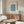 May include: Framed botanical poster with blue marigold pattern design displayed above wooden sideboard in modern living room. White textured chair and glass-top coffee table visible. Minimalist home decor styling with neutral walls and natural lighting.