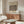 May include: Framed vintage botanical poster with honeysuckle pattern in muted greens and pinks displayed on wooden sideboard. Modern living room setting with cream upholstered chair, glass oval coffee table, and minimalist brass floor lamp. Warm natural lighting creates soft shadows on neutral walls.