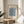 May include: Framed botanical art print featuring William Morris Marigold pattern in soft blue and cream tones, displayed on light wooden round side table next to beige upholstered chair. Natural sunlight streams across neutral-toned room with plant leaves visible in corner. Print shows intricate floral and leaf motifs in decorative Arts and Crafts style within thin gold frame.