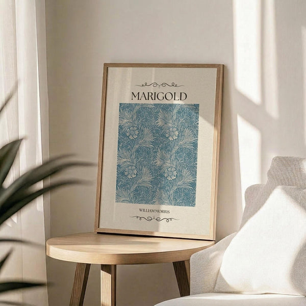 May include: Framed botanical art print featuring William Morris Marigold pattern in soft blue and cream tones, displayed on light wooden round side table next to beige upholstered chair. Natural sunlight streams across neutral-toned room with plant leaves visible in corner. Print shows intricate floral and leaf motifs in decorative Arts and Crafts style within thin gold frame.