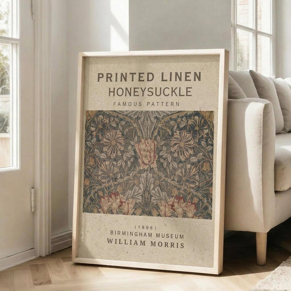 May include: Framed vintage botanical poster leaning against white wall next to beige upholstered chair. Print features intricate floral pattern with honeysuckle blooms in muted green, pink and brown tones on cream background. Text reads "Printed Linen Honeysuckle Famous Pattern" with museum attribution. Natural light streams through tall windows creating soft shadows on hardwood floor.