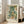 May include: Framed botanical art print leaning against white wall in bright living room. Light wood frame contains vintage-style floral pattern with sage green leaves, cream and peach flowers, and small berries. Text reads "WILLIAM MORRIS Exhibition Art - Floral Fantasy" below ornate design. Beige sofa and large windows visible in background, natural lighting creates soft shadows on hardwood floor.