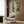 May include: Framed Studio Ghibli My Neighbor Totoro movie poster hanging on wood-paneled wall above beige upholstered armchair with geometric patterned pillow. Poster shows Totoro characters in lush green forest scene with Japanese text. Modern reading nook with brass floor lamp, side table with open book, and natural lighting from window with curtains.