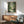 May include: Framed My Neighbor Totoro poster displayed above a wooden sideboard with white ceramic lamp. The green forest artwork shows the beloved character sitting by a stream with an umbrella, surrounded by lush trees. Mid-century modern living room setting with sage green armchair, round coffee table, and natural lighting casting shadows on white wall.