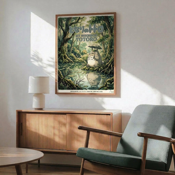 May include: Framed My Neighbor Totoro poster displayed above a wooden sideboard with white ceramic lamp. The green forest artwork shows the beloved character sitting by a stream with an umbrella, surrounded by lush trees. Mid-century modern living room setting with sage green armchair, round coffee table, and natural lighting casting shadows on white wall.