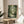 May include: Framed poster displayed on wooden round side table next to beige upholstered chair in sunlit room with white walls and plant. Artwork shows forest scene with large gray creature holding umbrella beside stream, surrounded by lush green trees and foliage in detailed illustrated style with Japanese text.