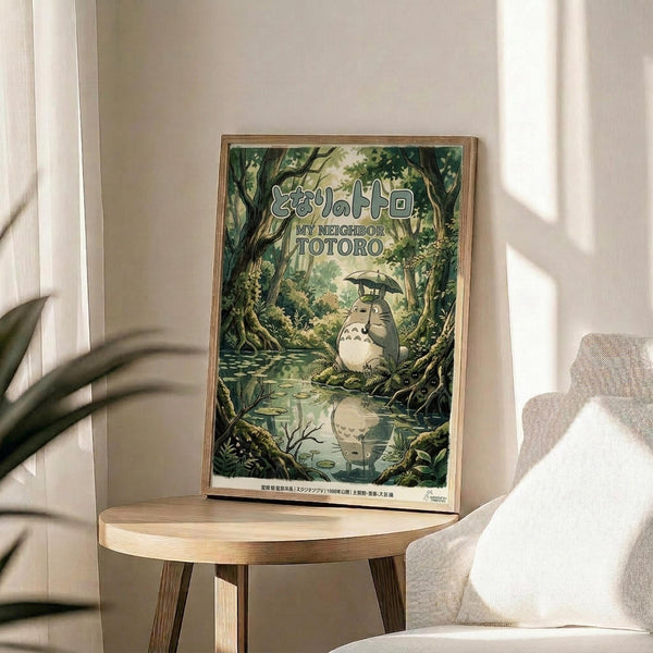 May include: Framed poster displayed on wooden round side table next to beige upholstered chair in sunlit room with white walls and plant. Artwork shows forest scene with large gray creature holding umbrella beside stream, surrounded by lush green trees and foliage in detailed illustrated style with Japanese text.
